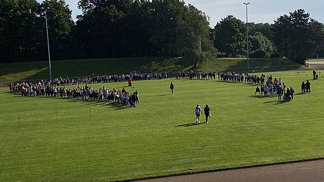 Auf der Wiese des Klever Stadions steht ein großer Kreis mit allen Kindern und Lehrenden der Karl-Leisner-Schule. 