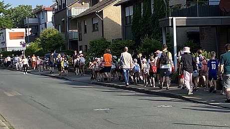 Auf dem Bild ist eine lange Schlange von Kindern und Betreuenden zu sehen, die über den Bürgersteig entlang der Straße laufen. 