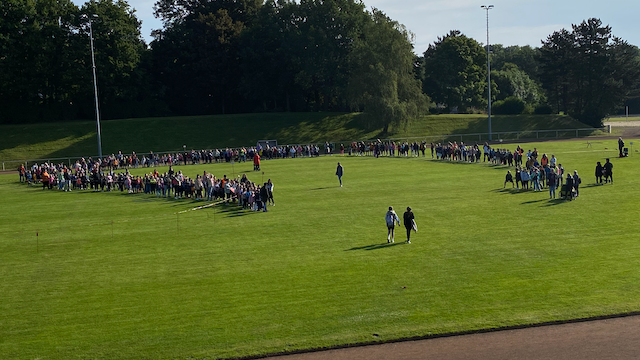 Auf dem Bild ist ein riesiger Kreis aus Kindern, Lehrenden und Eltern der Karl-Leisner-Schule auf der Stadionwiese zu sehen. 